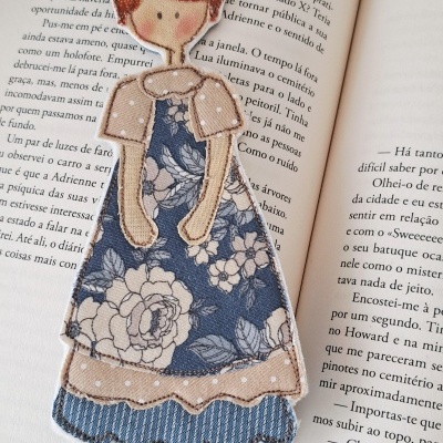 Marcador de página em tecido com figura de menina em vestido azul floral e cabelo castanho, colocado num livro aberto.