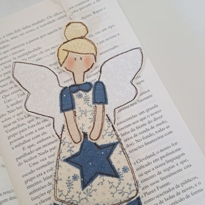 Marcador de livros de tecido com anjo em tons de azul, bege e branco sobre livro aberto