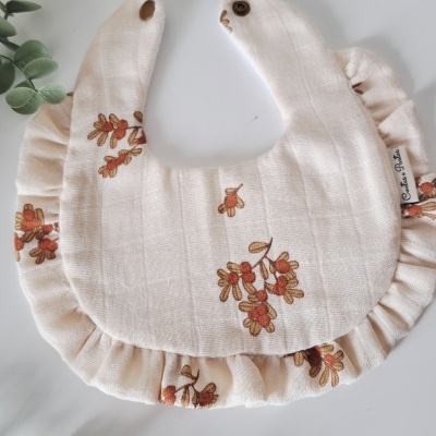 Babete de bebé cru com padrão floral e folhos rodeado por folhas verdes