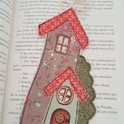 Marcador de livros em tecido com design de casinhas e padrões florais colocado sobre livro aberto.