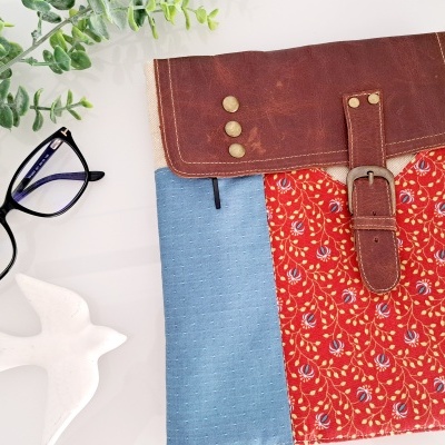 Bolsa de tecido azul e vermelho com fecho em couro castanho, óculos pretos, figura de pássaro branca e plantas verdes