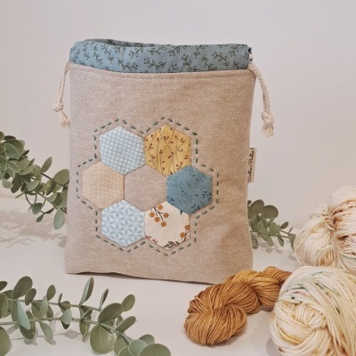 Saquinho tecido com padrão hexagonal e cordão, em ambiente com lã e ramos verdes