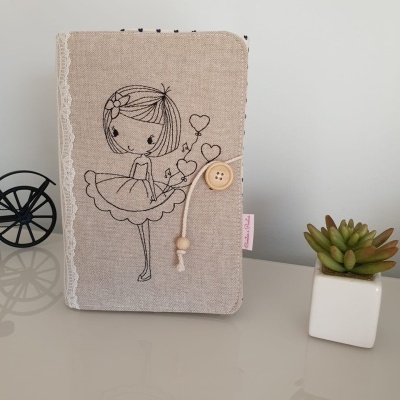 Capa de tecido bege com desenho de menina e coração, botão e cordão, renda branca, superficie espelhada com vaso branco e planta suculenta