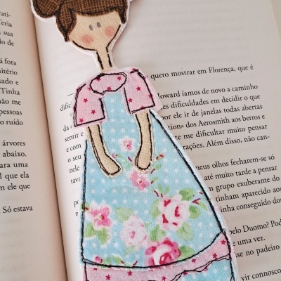 Marcador de livros em formato de menina com vestido azul floral e rosa com estrelas em tecido sobre livro aberto