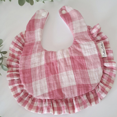 Babete para bebé com padrão xadrez rosa e branco, com bordas plissadas, colocado sobre uma mesa branca cercada por folhagem verde.