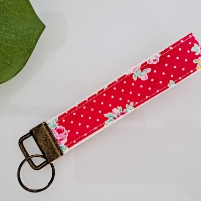 Chaveiro tecido vermelho com pintas brancas e flores rosa ao lado de folha verde