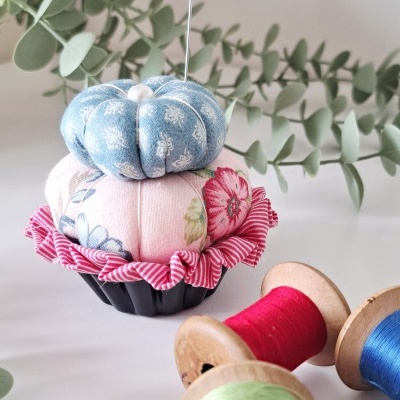 Alfineteiro de tecido em forma de bolo com cores azul, rosa e padrão floral, com carretéis de linha colorida e folhagem verde ao fundo