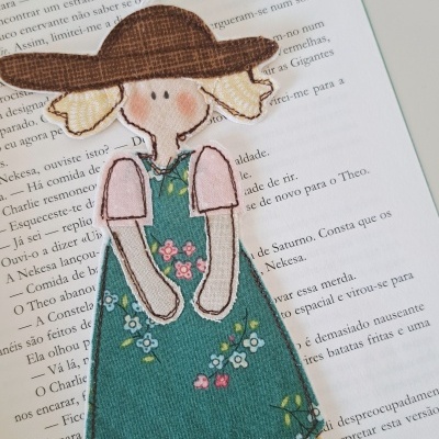 Marcador de página de tecido em forma de boneca com vestido verde e chapéu castanho sobre livro aberto