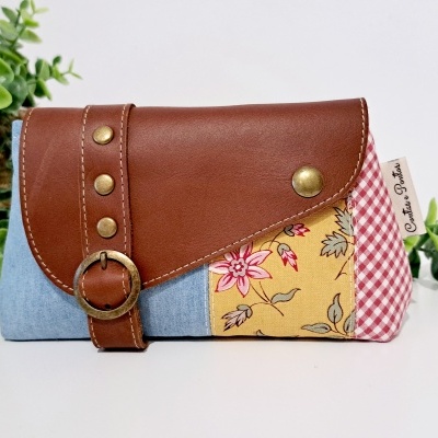 Carteira multicolorida com couro castanho, tecido azul e tecido amarelo floral, com padrão xadrez vermelho e branco, etiquetada 'Coisas Da Júlia', sobre fundo branco com plantas verdes.