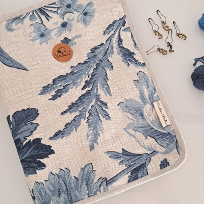 Estojo de tecido bege com padrão floral azul, etiqueta e botão handmade, junto a novelos de lã azul e alfinetes de número.