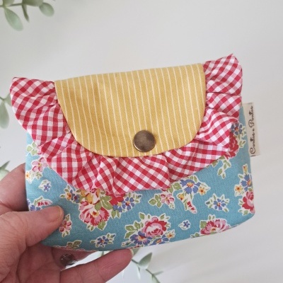 Carteira de tecido azul com estampado floral, aba amarela riscada, dobra em tecido xadrez vermelho e branco e botão de pressão