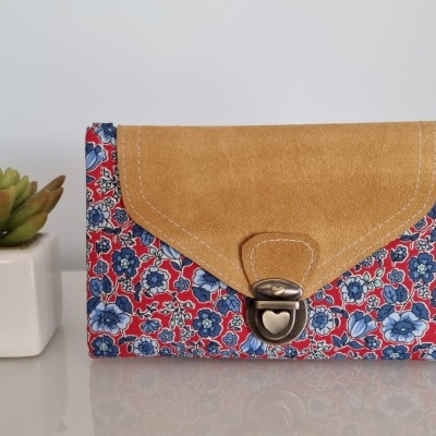 Carteira com tecido floral vermelho e azul e aba castanha com fecho metálico em forma de coração ao lado de planta suculenta em vaso branco