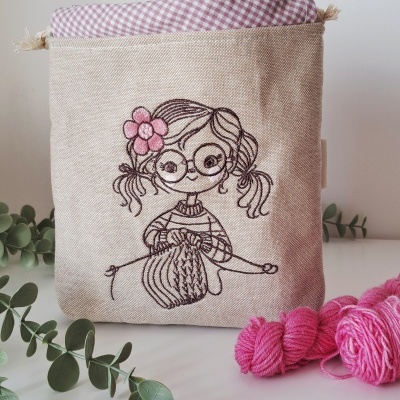 Saco de tecido bege com bordado de menina a tricotar e forro xadrez rosa