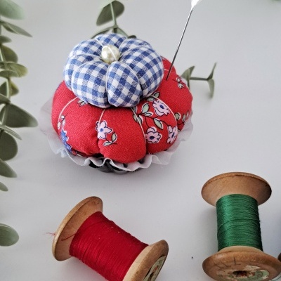 Alfineteiro com tecido xadrez azul e vermelho com flores, com linhas vermelha e verde e folhas verdes ao redor