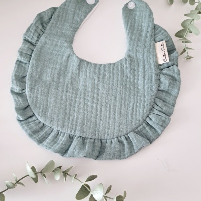 Babete azul esverdeado com franzidos e fecho branco sobre fundo branco e folhas verdes