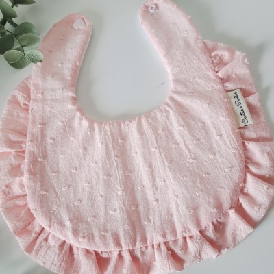 Babete cor-de-rosa claro com textura, ruffles, botão de pressão e etiqueta, sobre superfície branca com ramos verdes