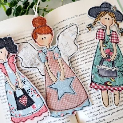 Três bonecas de tecido coloridas com bordados e aplicações, sobre um livro aberto.