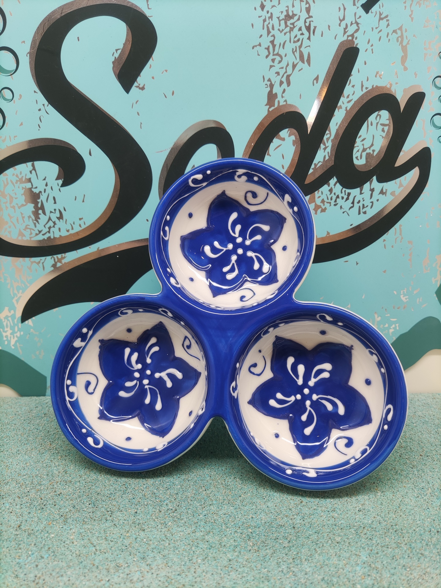 Conjunto de três pratos cerâmicos azuis e brancos com flores azuis, fundos azul e verde, palavra 'Soda' ao fundo