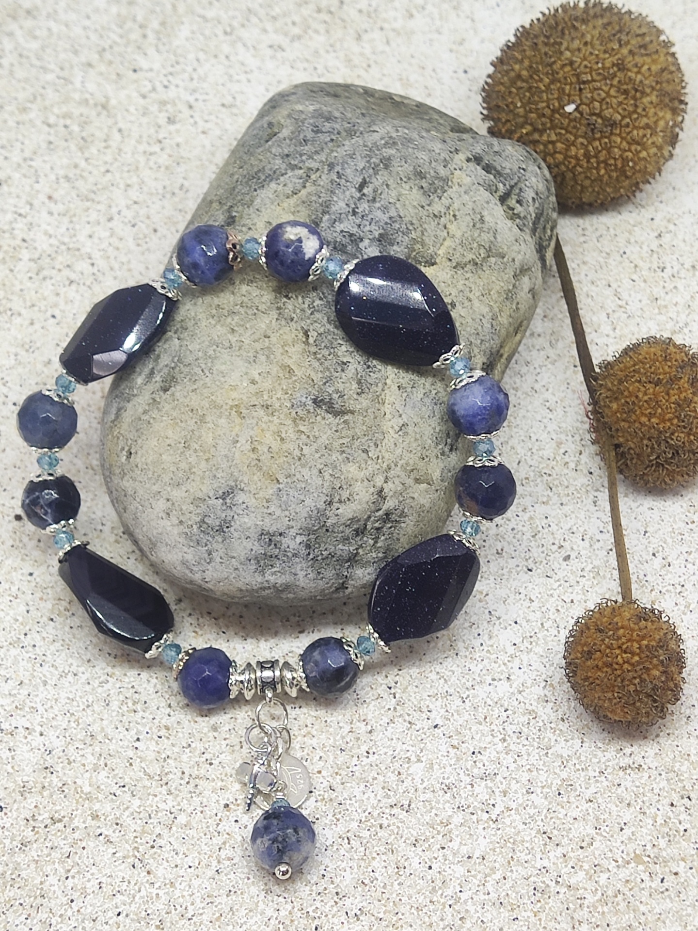 Pulseira azul com pingente sobre pedra cinzenta e fundo de areia clara com ramos secos