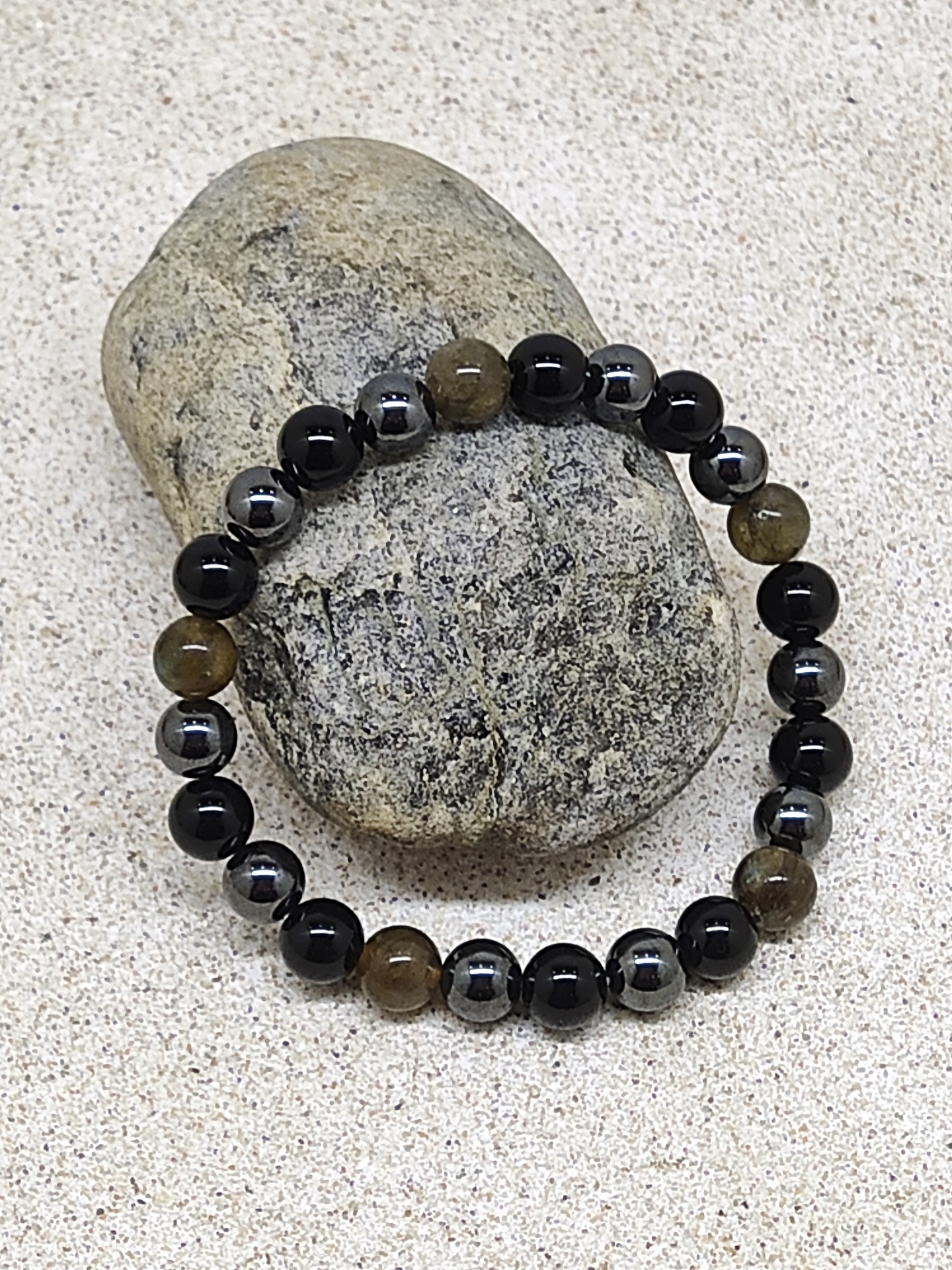 Pulseira elástica com contas pretas, cinzentas metálicas e castanhas sobre pedra cinzenta em areia clara