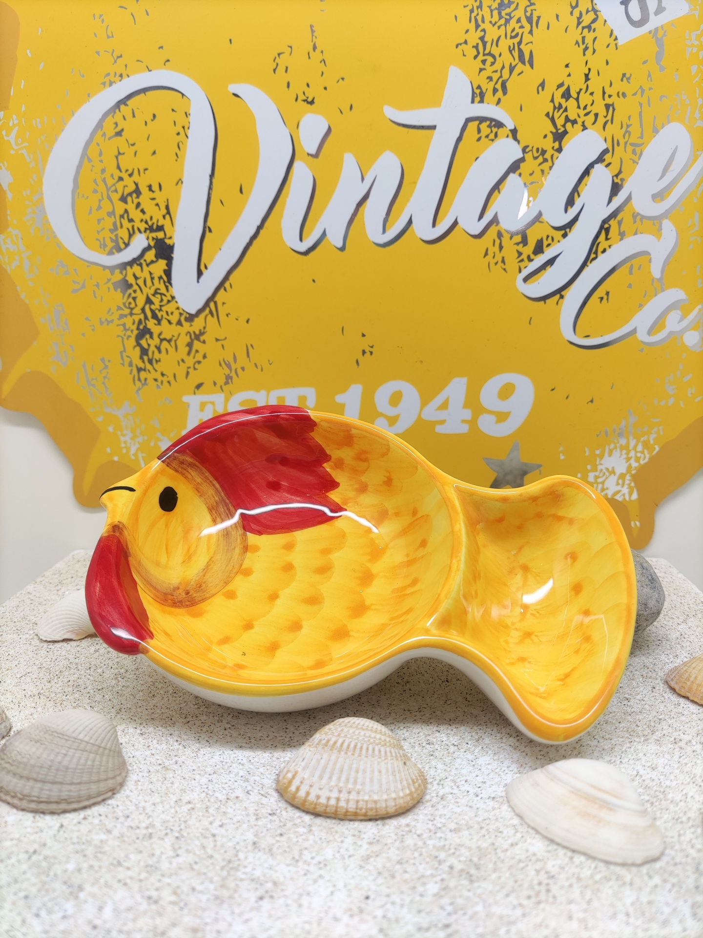 Prato em forma de peixe amarelo com detalhes vermelhos sobre areia com conchas e cartaz amarelo ao fundo