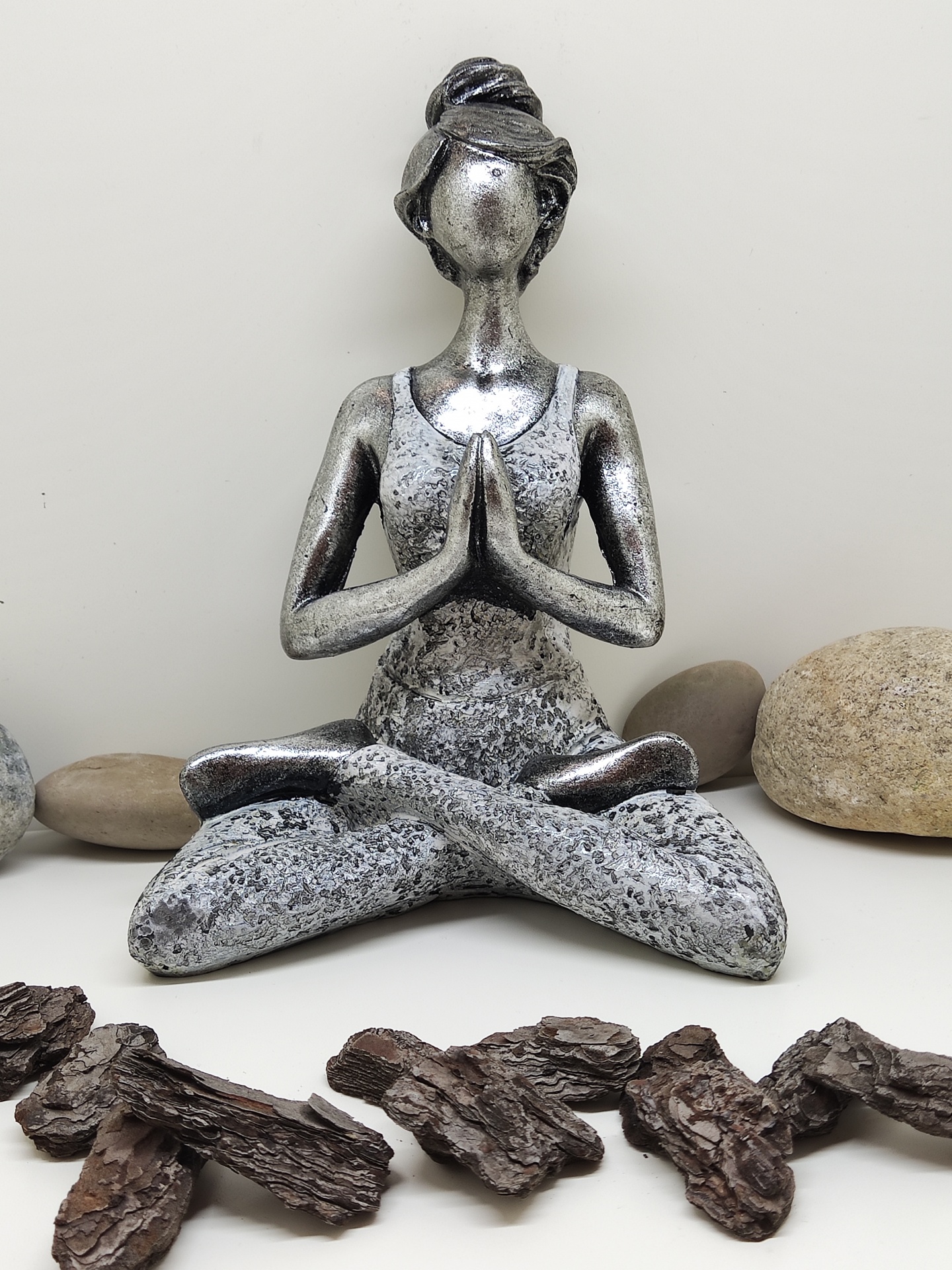 Estátua metálica prateada de mulher em posição de meditação com pedras e cascas ao redor