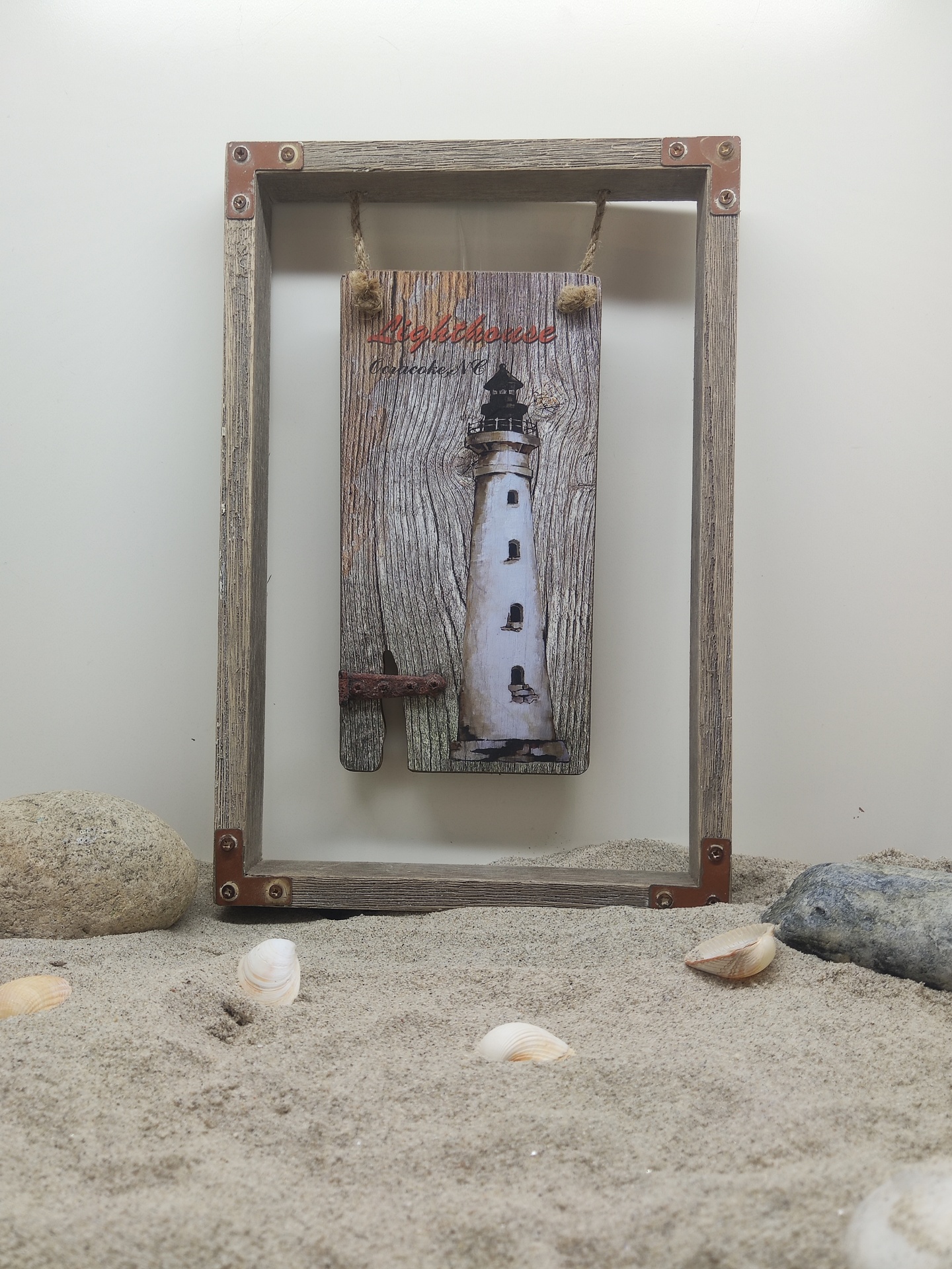 Quadro decorativo com farol e texto Algarve Portugal em madeira rústica pendurada dentro de moldura de madeira sobre areia com conchas e pedras