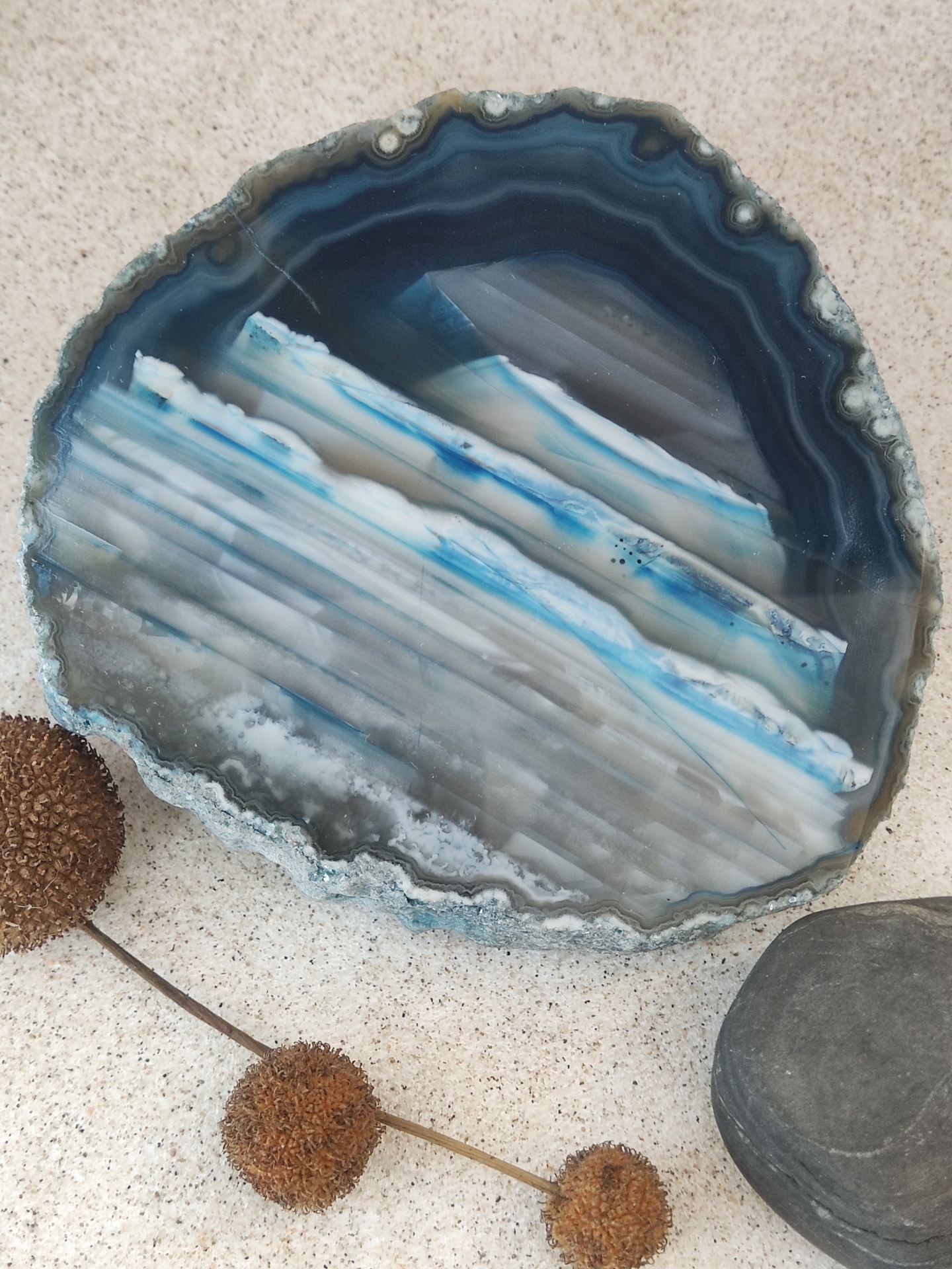 Fatia de ágata azul sobre superfície clara com flores secas e pedra cinza