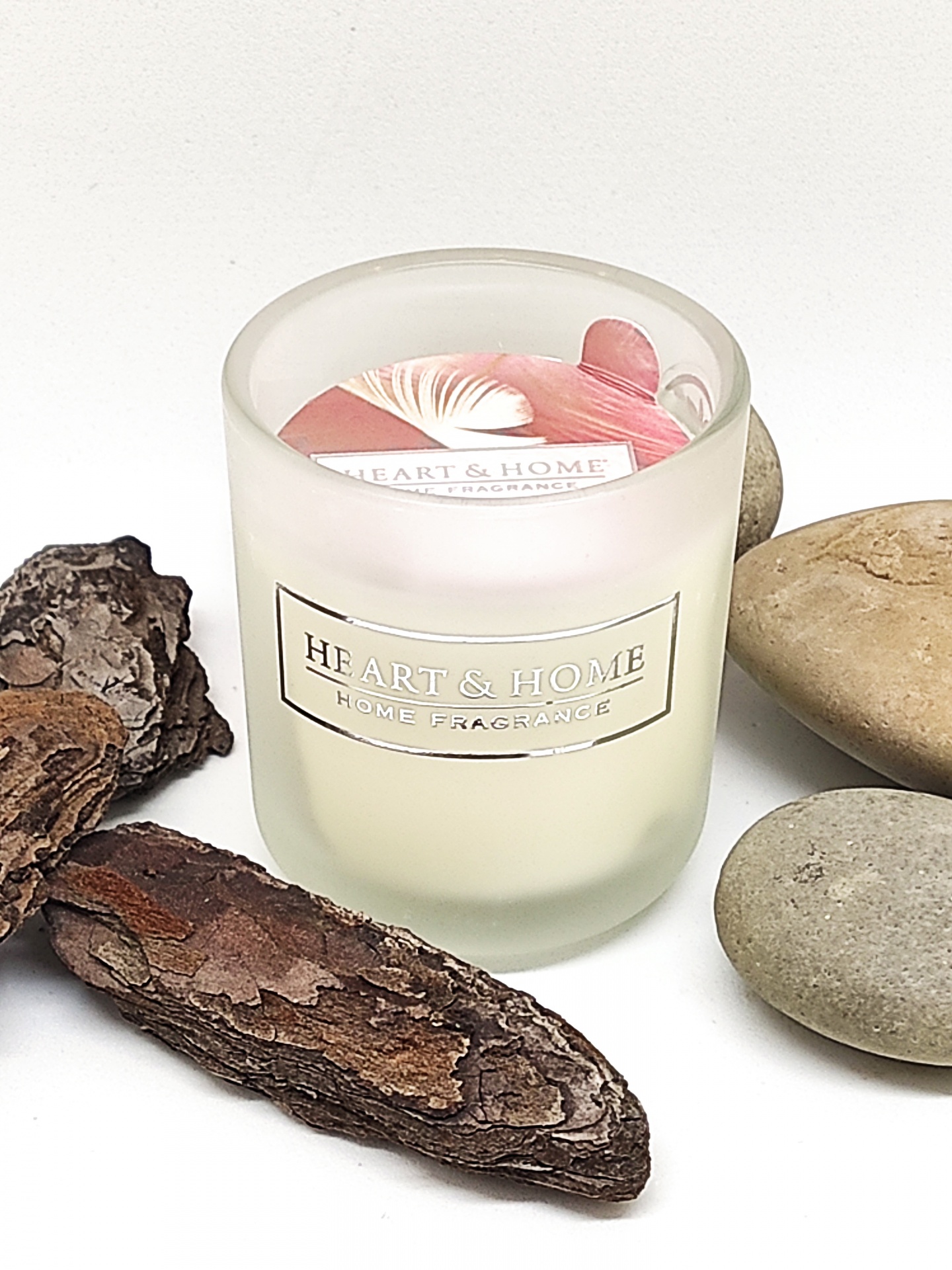 Vela aromática HEART & HOME em copo de vidro fosco com madeira e pedras ao redor em fundo branco