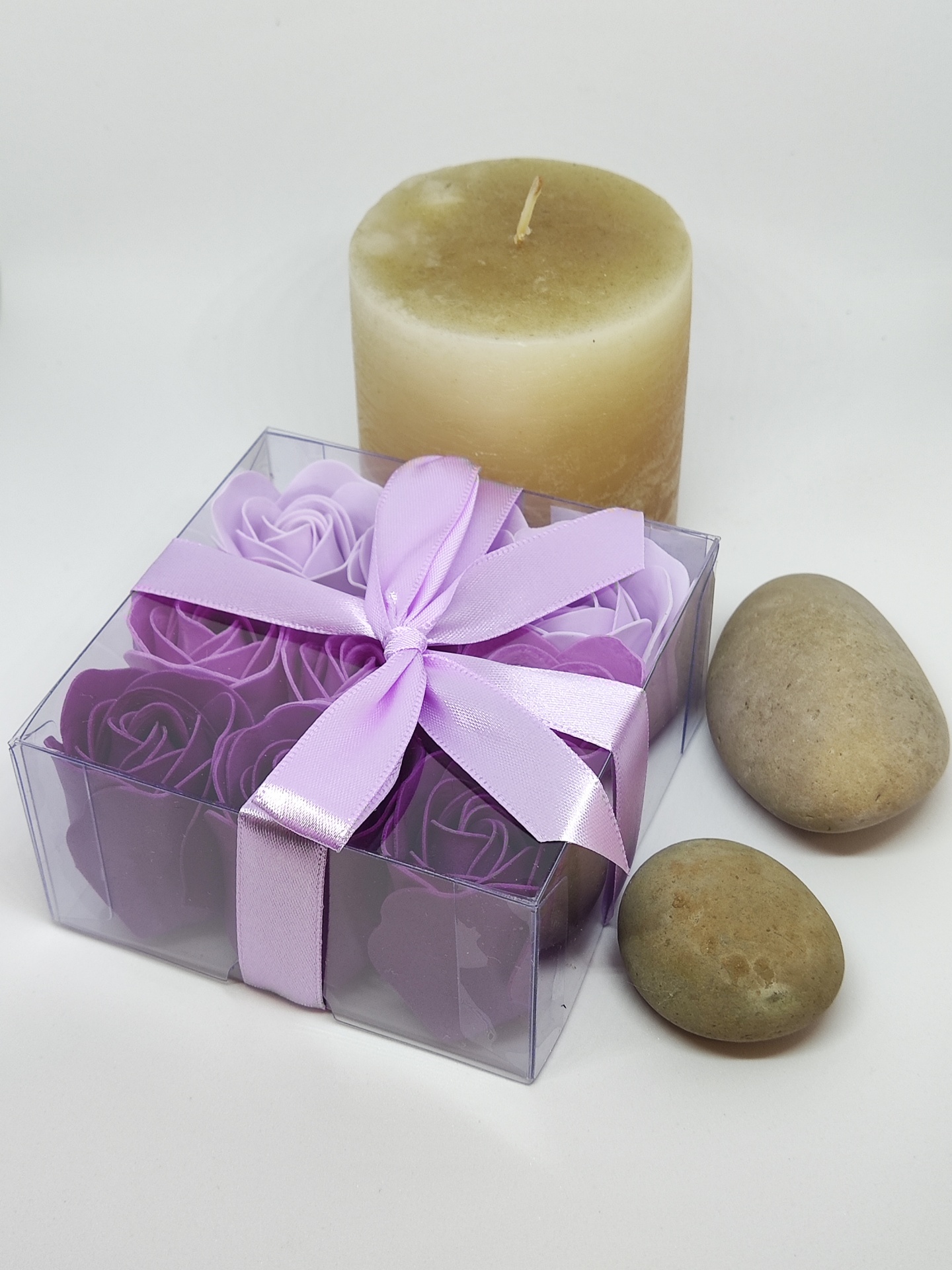 Caixa transparente com flores de sabonete lilás, vela creme e pedras ovais num fundo branco