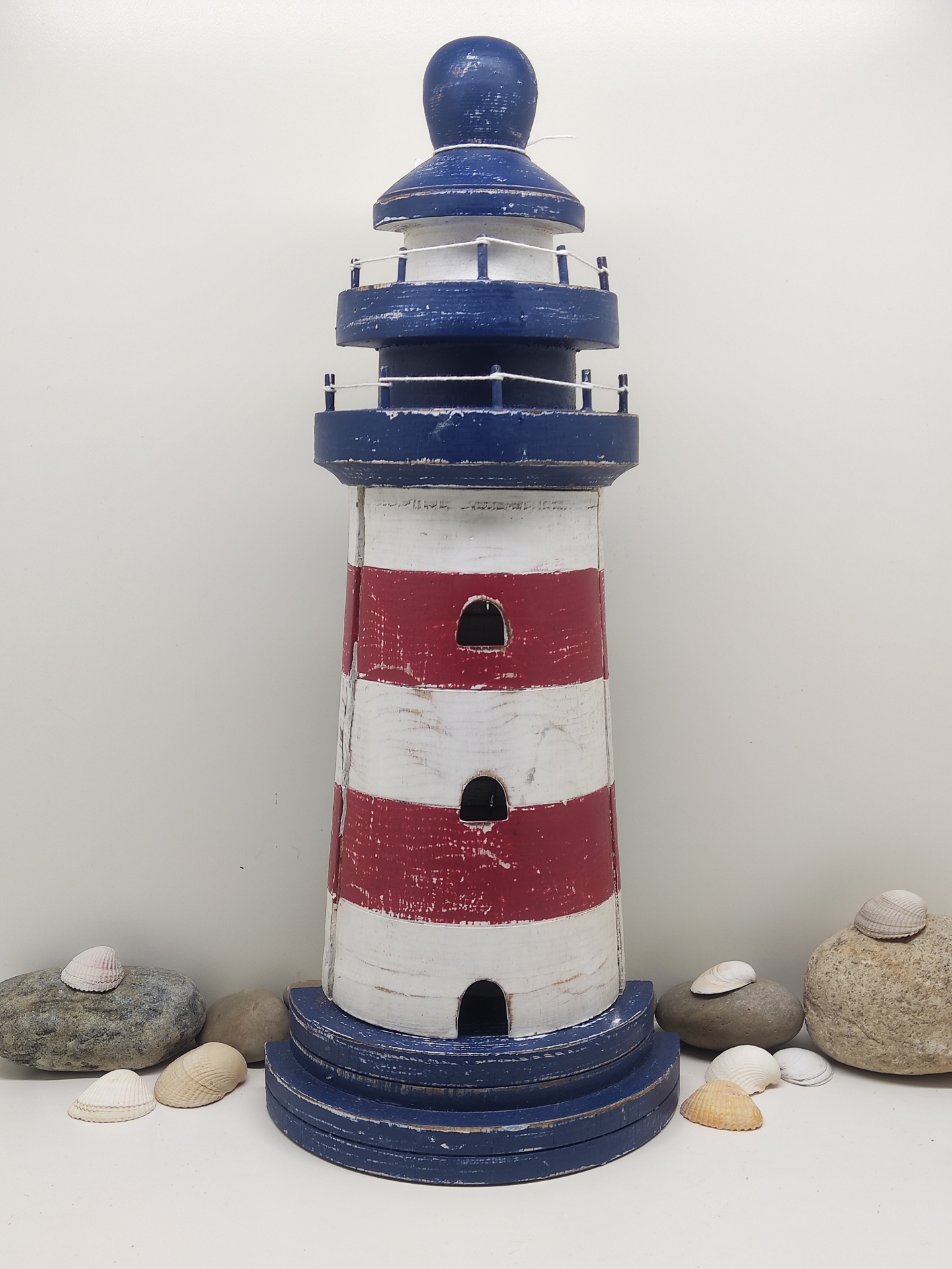 Farol decorativo de madeira com riscas vermelhas e brancas e cúpula azul rodeado por pedras e conchas