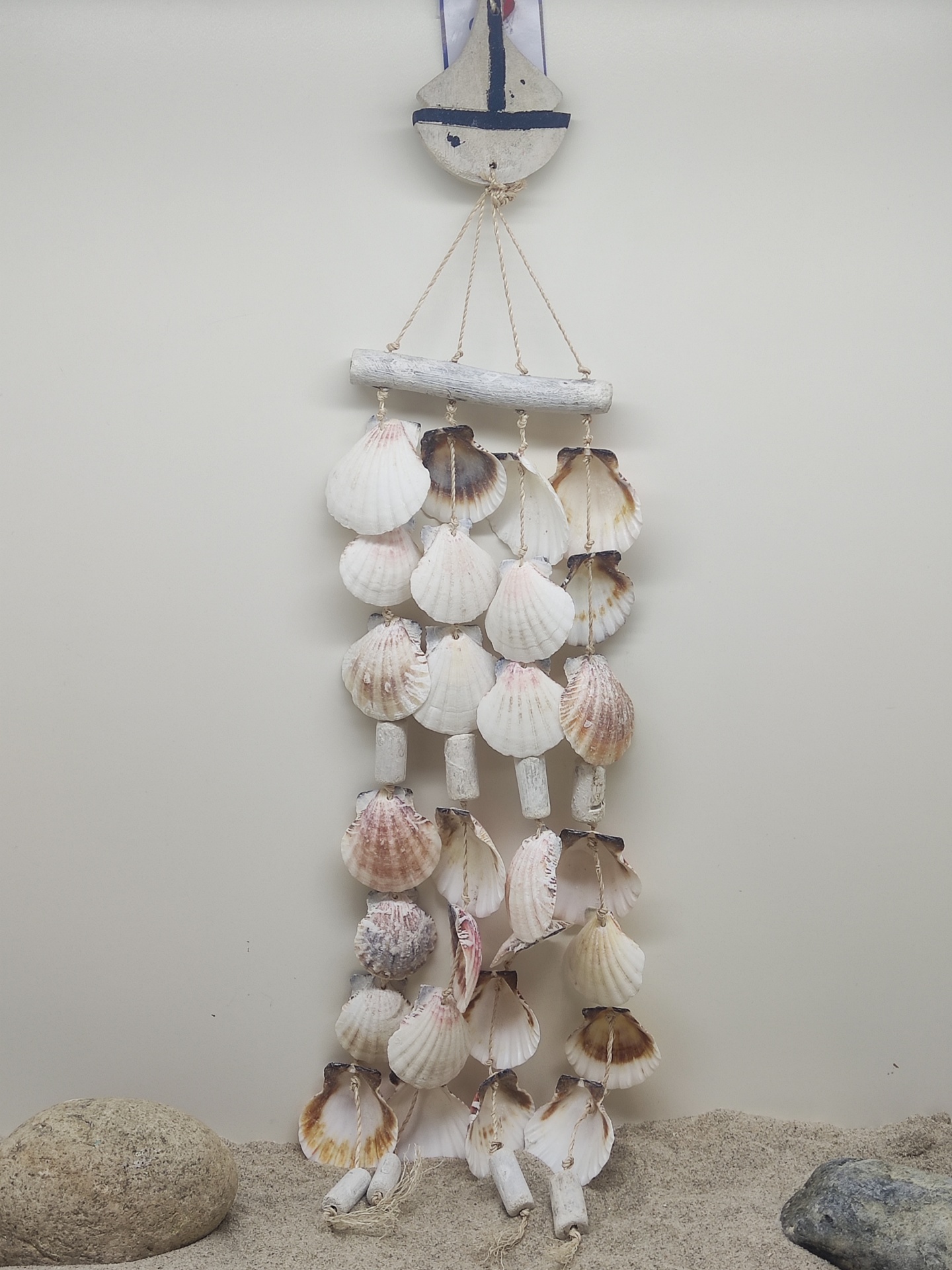 Móvel pendente feito de conchas naturais em tons claros e peças de madeira branca com detalhes azuis, pendurado numa parede branca, sobre areia e pedras.