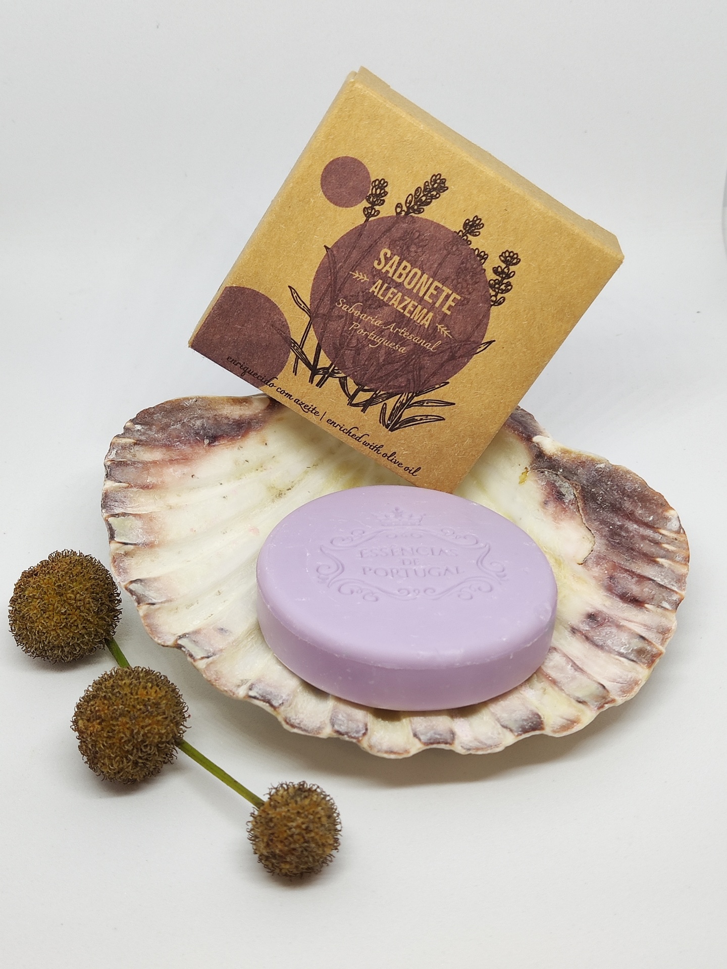 Sabonete lilás dentro de concha com embalagem castanha e flores secas ao lado.