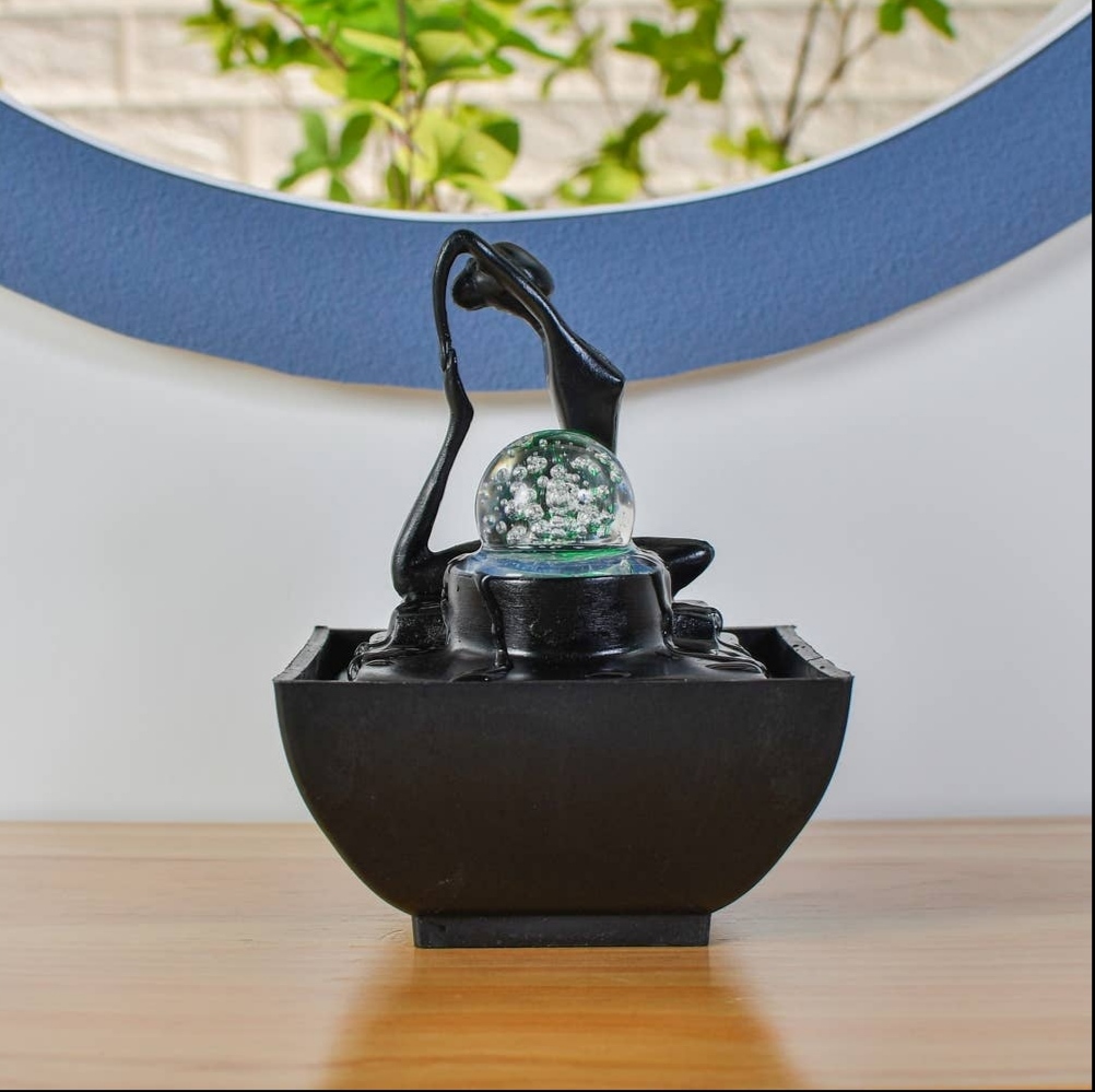 Fonte decorativa preta com bola de vidro e figura abstrata, em superfície de madeira, frente a parede branca e espelho azul.