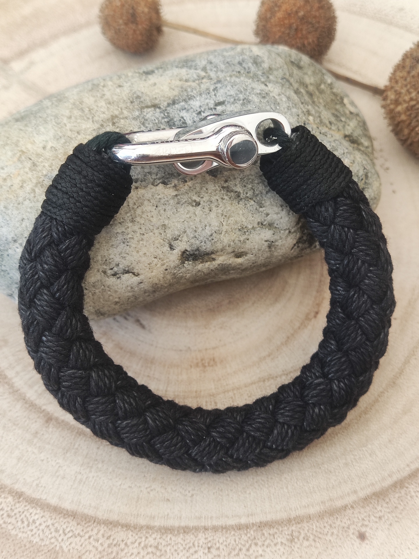 Pulseira de corda preta trançada com fecho metálico prateado sobre pedra e fundo de madeira