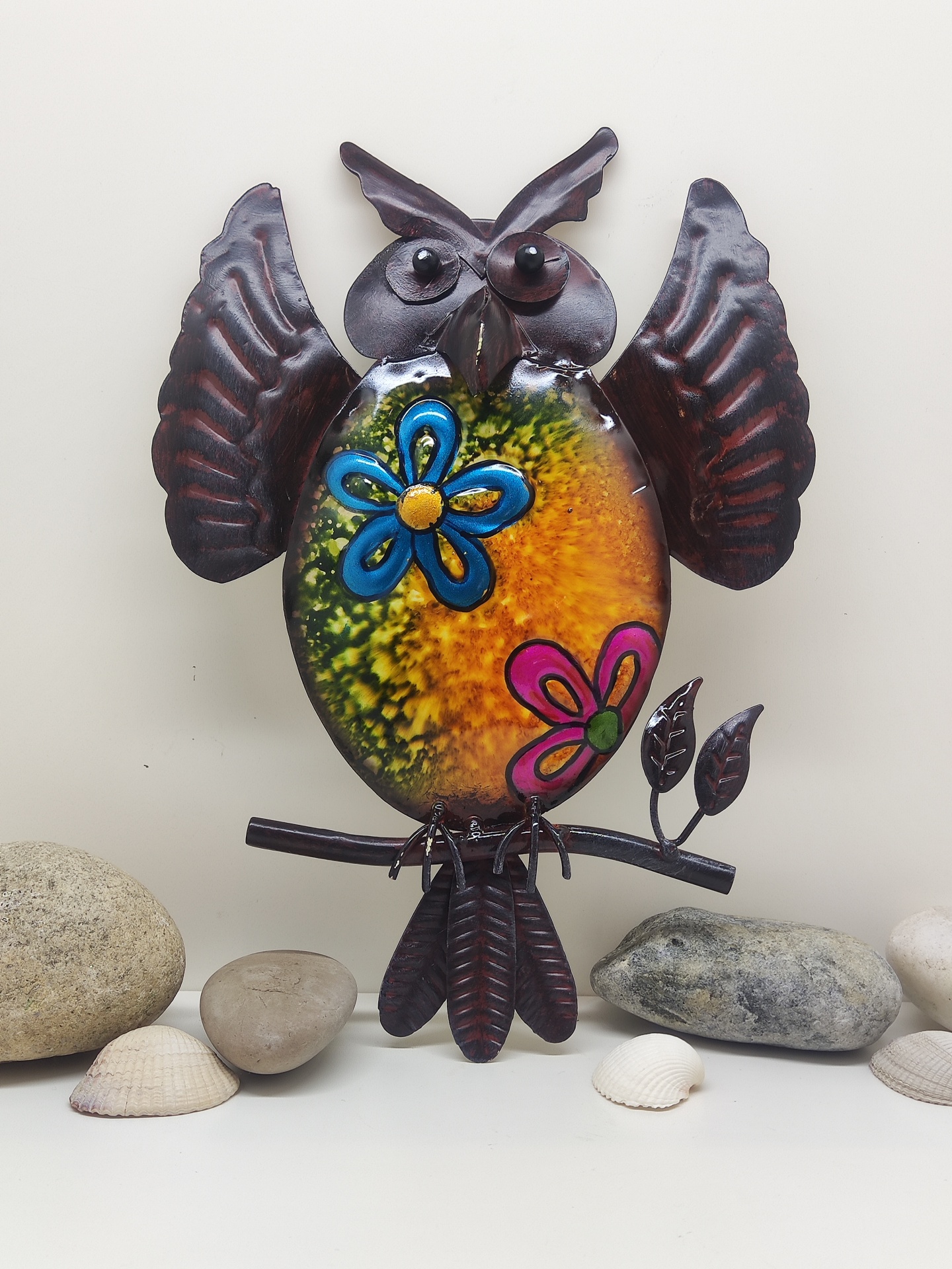 Estatueta de coruja decorativa em metal com padrão colorido e flores, rodeada por pedras e conchas.