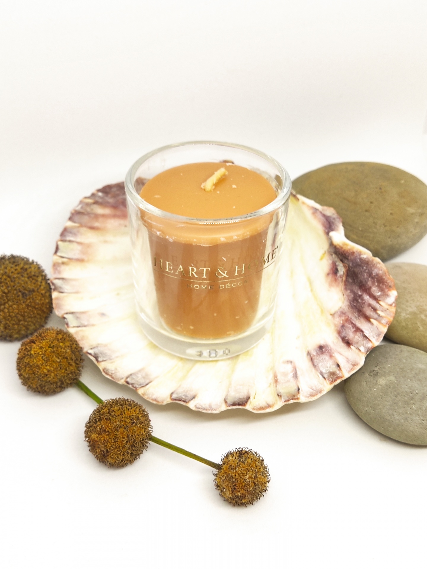 Vela laranja em copo de vidro sobre concha rodeada de pedras e flores secas.