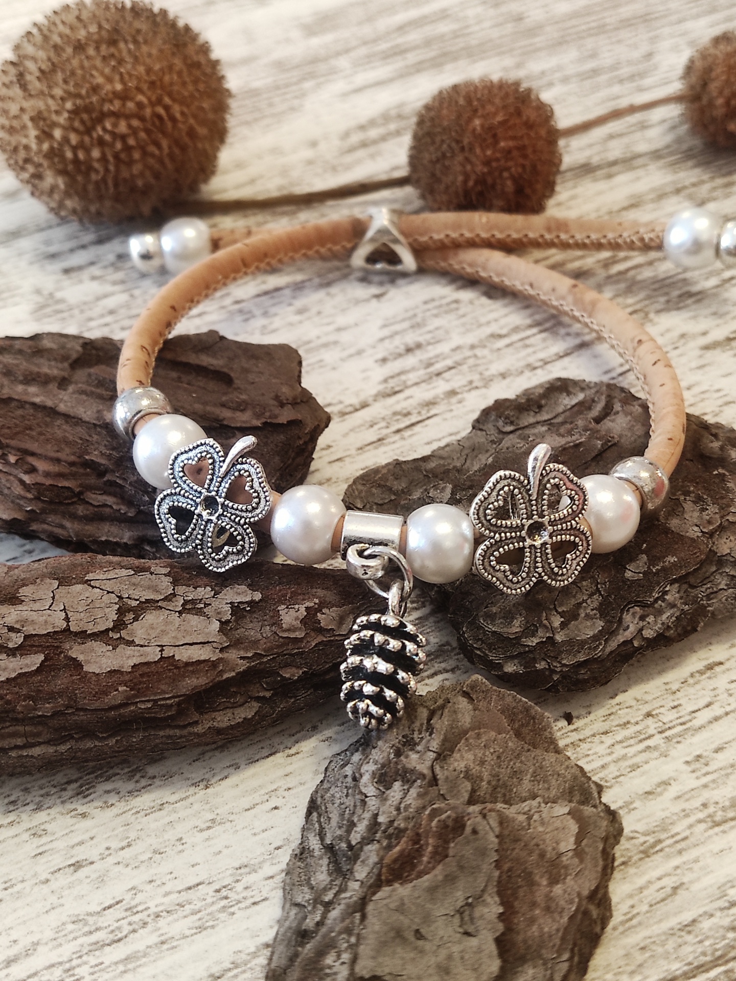 Pulseira em cortiça com contas brancas, ornamentos florais metálicos e pendente de pinha sobre fundo de madeira e casca