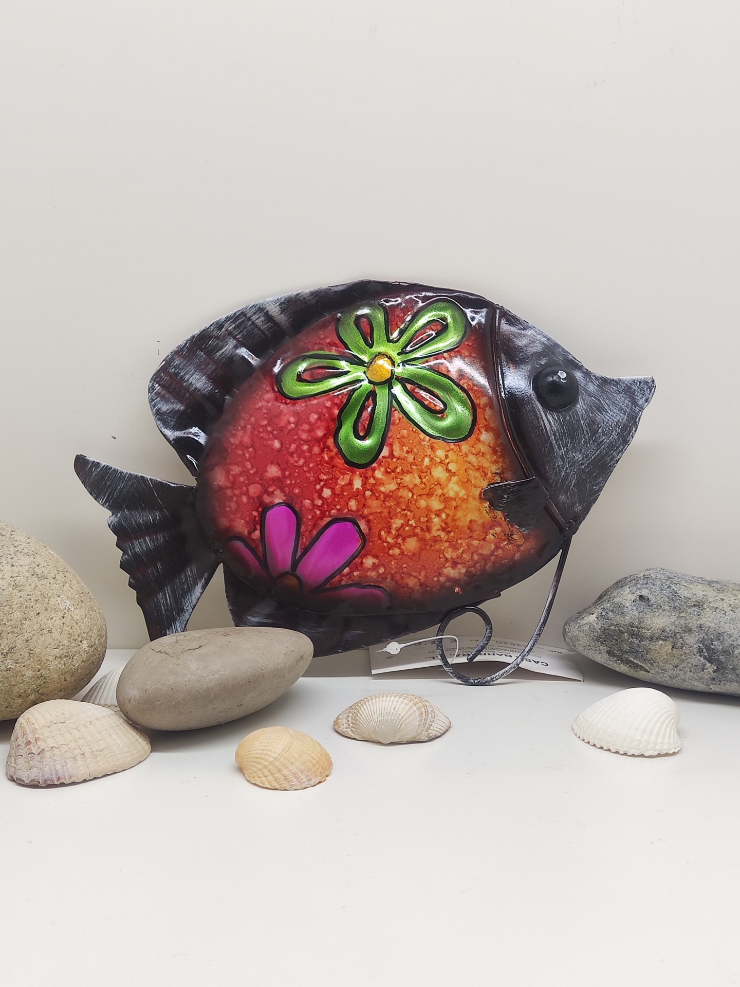 Peixe decorativo metálico colorido com padrão de flores entre pedras e conchas