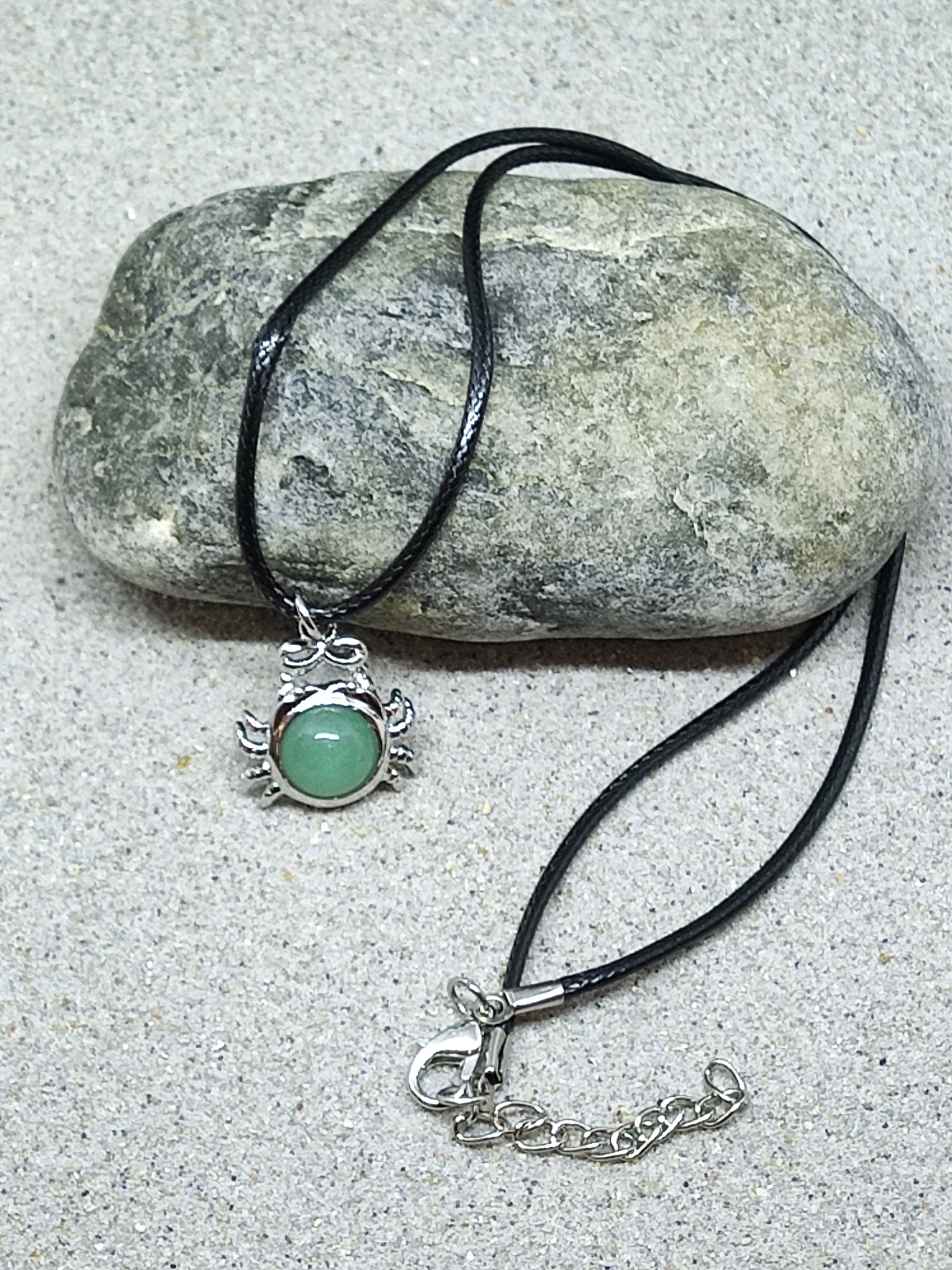 Colar com pendente de caranguejo prateado e pedra verde em cordão preto sobre pedra e areia