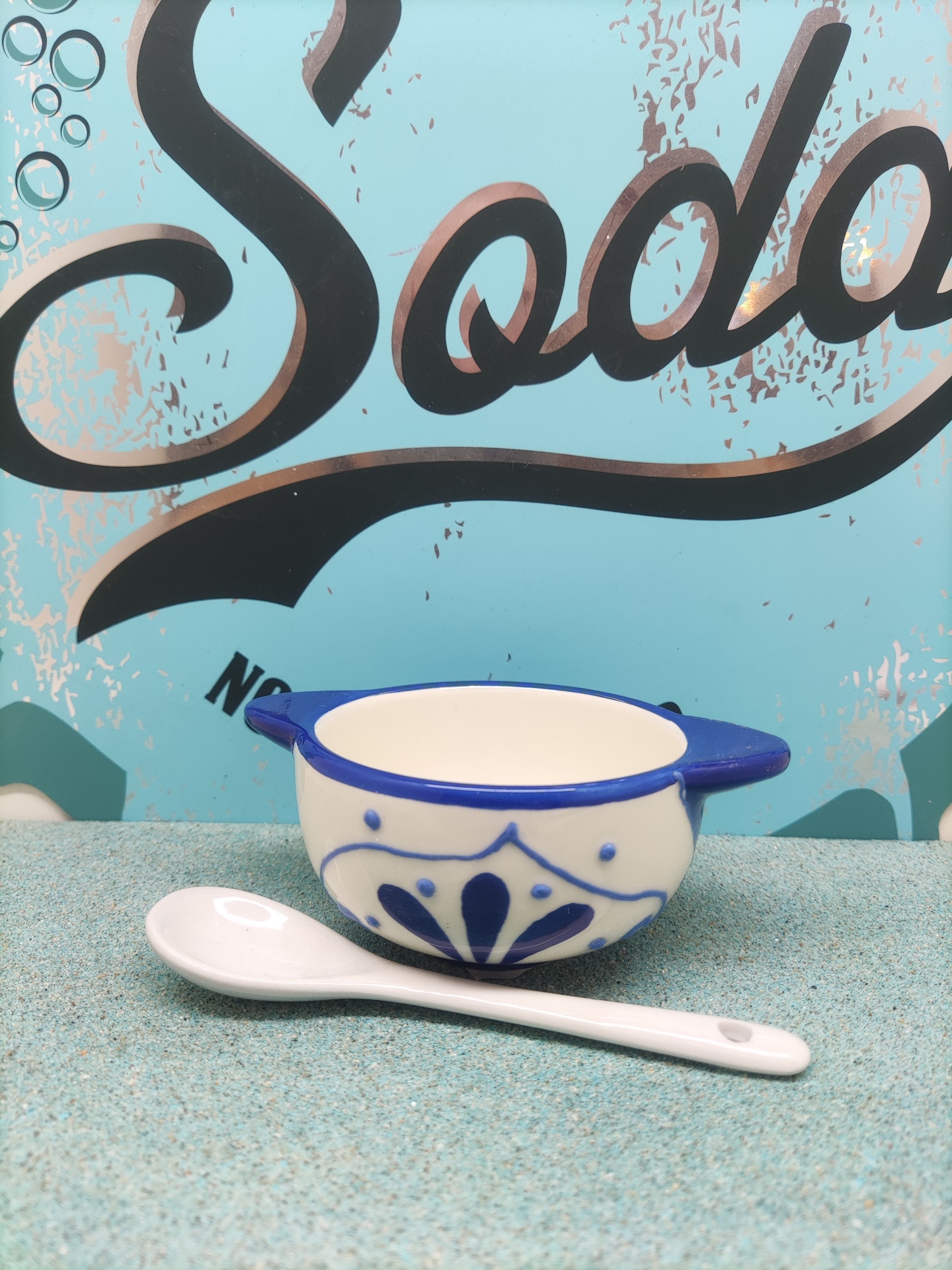 Tigela de cerâmica branca e azul com colher branca em superfície azul clara e fundo com texto 'Soda'