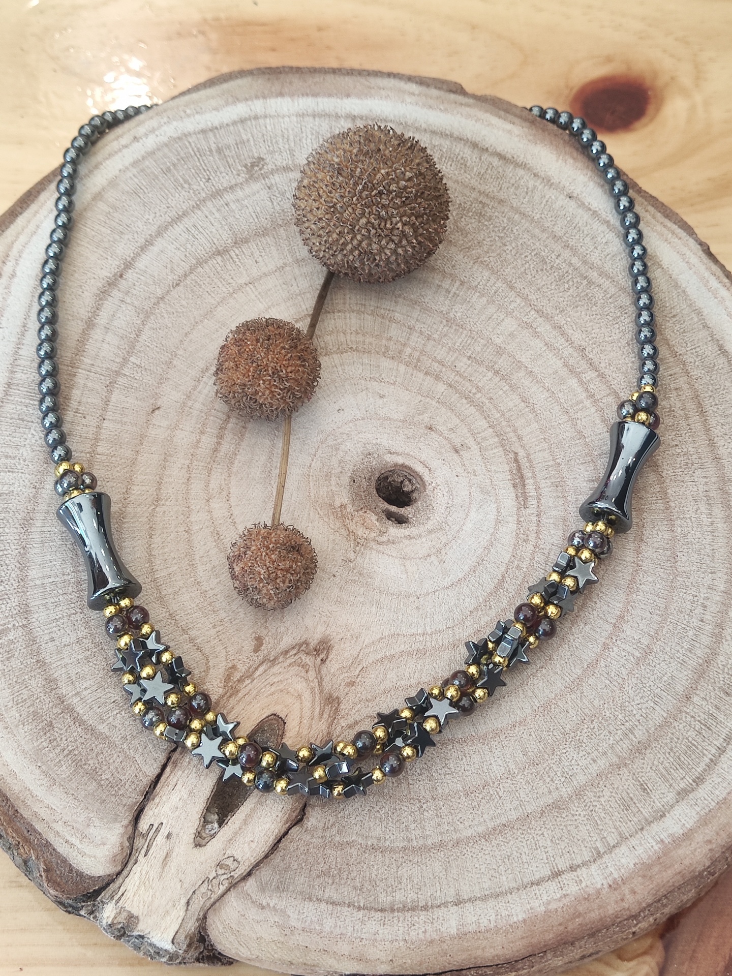 Colar com contas metálicas cinzentas e douradas sobre base de madeira com sementes secas