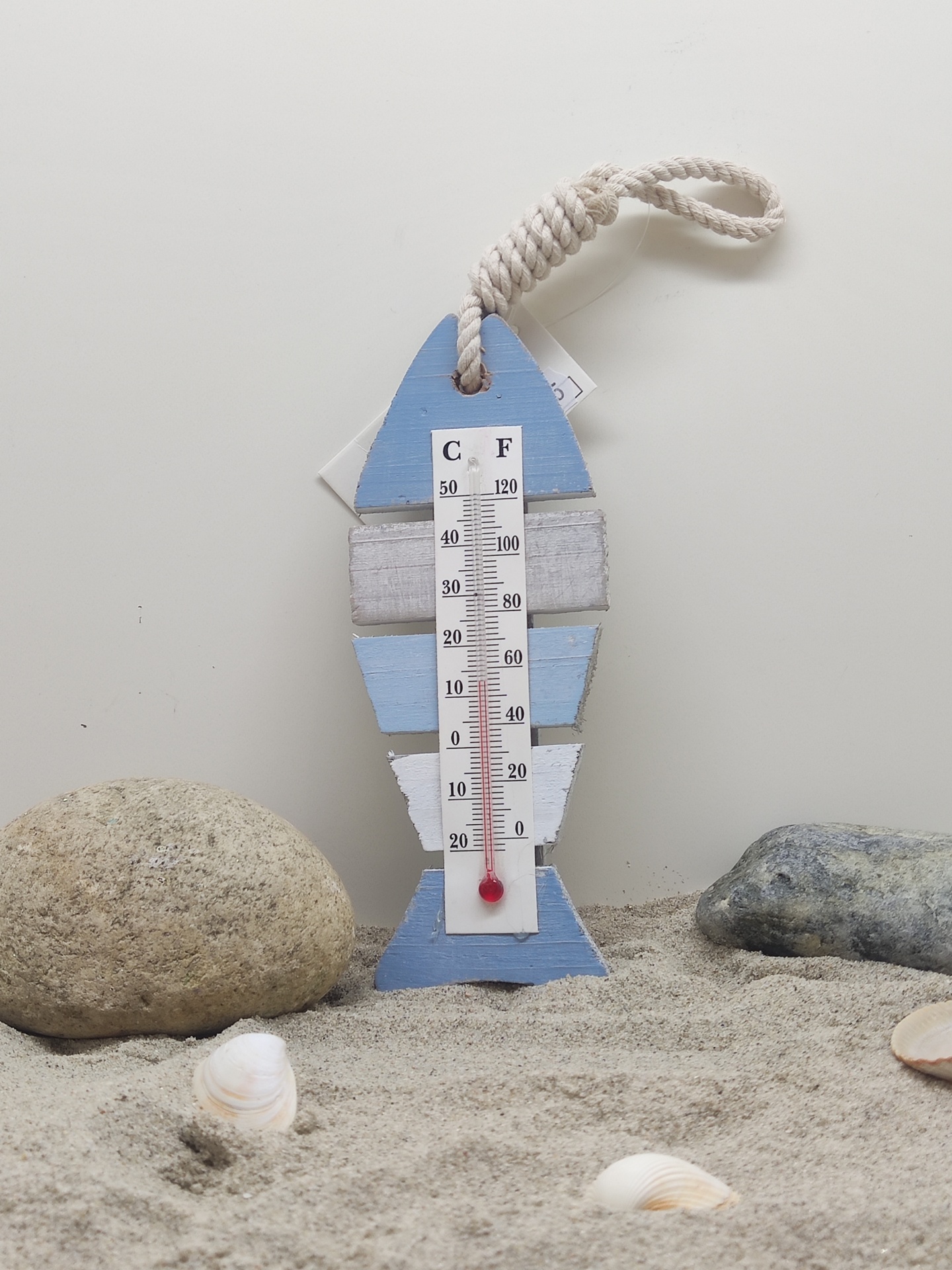 Termómetro decorativo em forma de peixe em madeira azul e branca com corda, na areia entre pedras e conchas