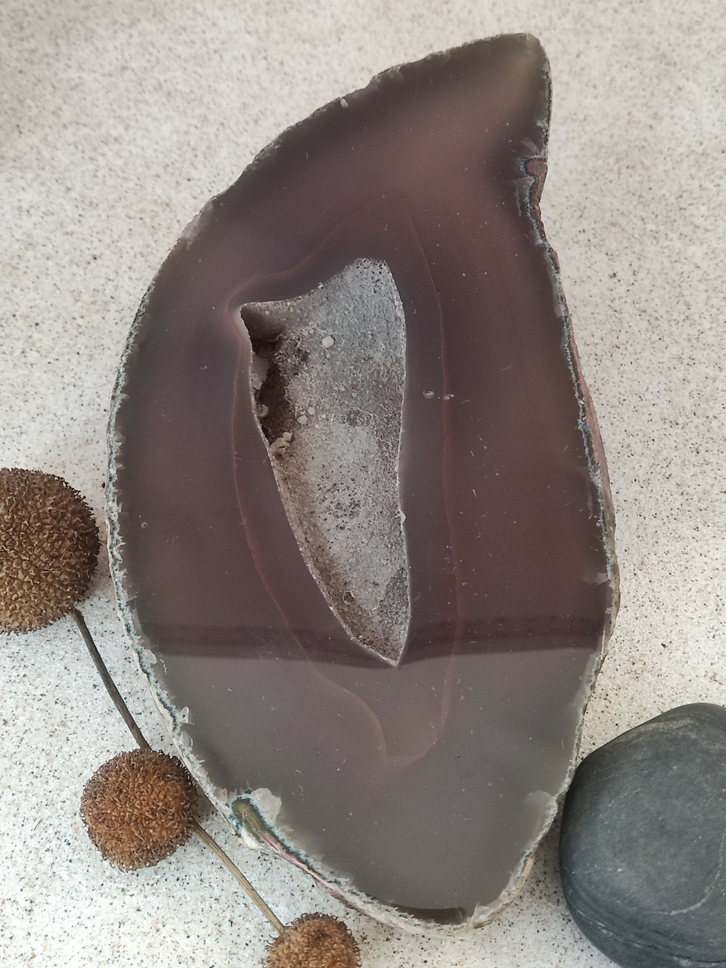 Peça decorativa de ágata castanha com cavidade, ao lado de pedras e plantas secas sobre superfície branca