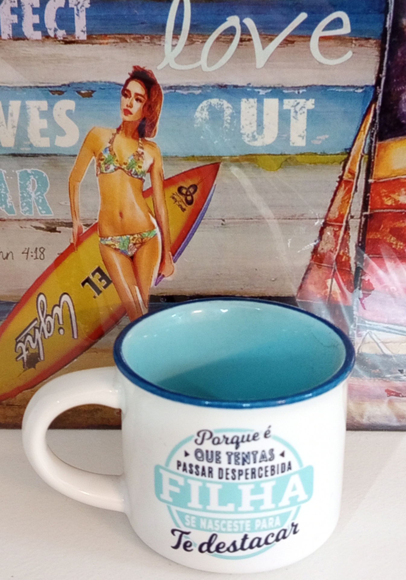Caneca branca com interior azul e texto em português, ao lado de imagem de mulher de fato de banho com prancha de surf.