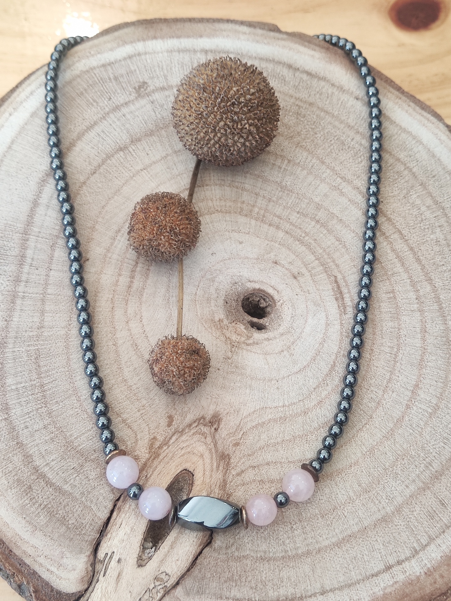 Colar com contas metálicas prateadas, contas rosa claro e conta preta oblonga sobre tábua de madeira clara