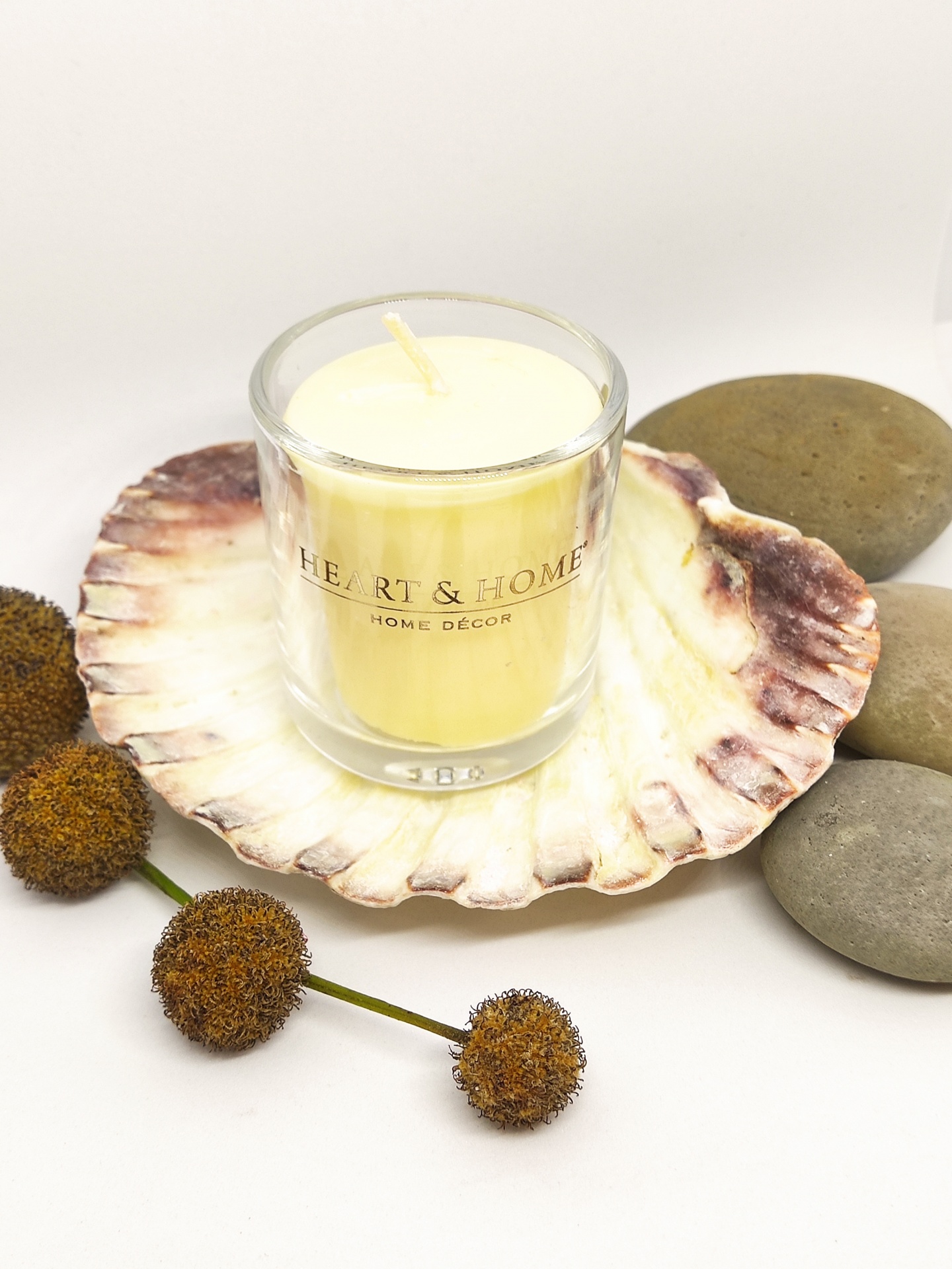 Vela amarela em copo de vidro sobre concha marrom e branca com pedras cinza e bolas secas castanhas