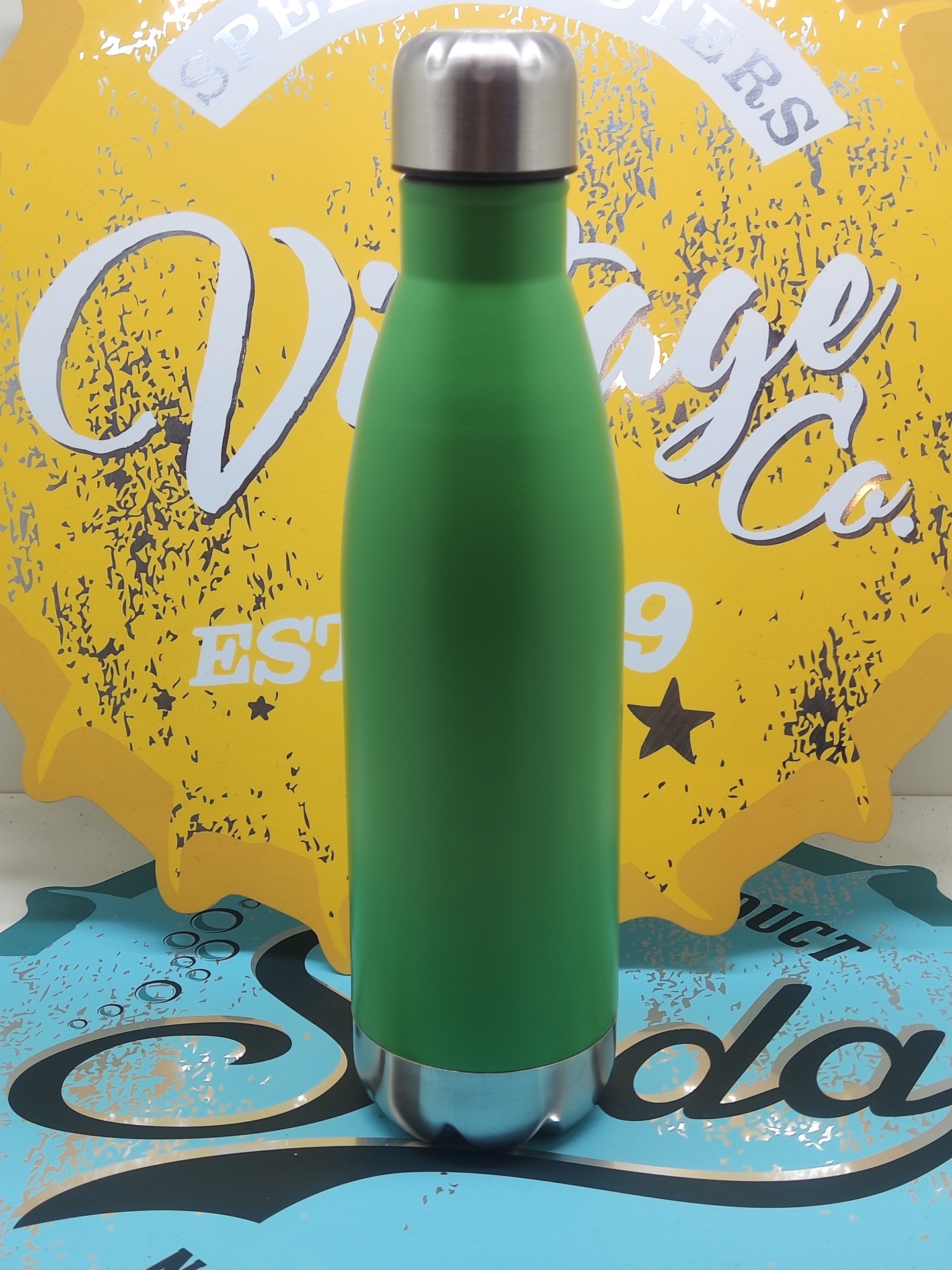 Garrafa térmica verde com tampa e base em aço inoxidável diante de fundo decorativo amarelo e azul