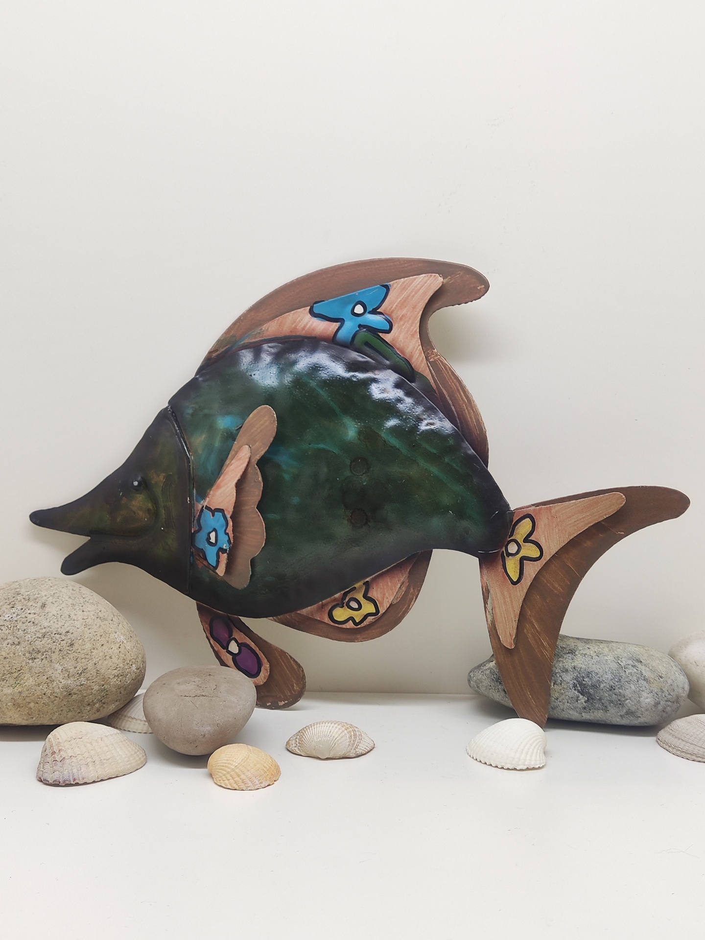 Peixe decorativo em madeira e metal com flores coloridas rodeado por pedras e conchas.