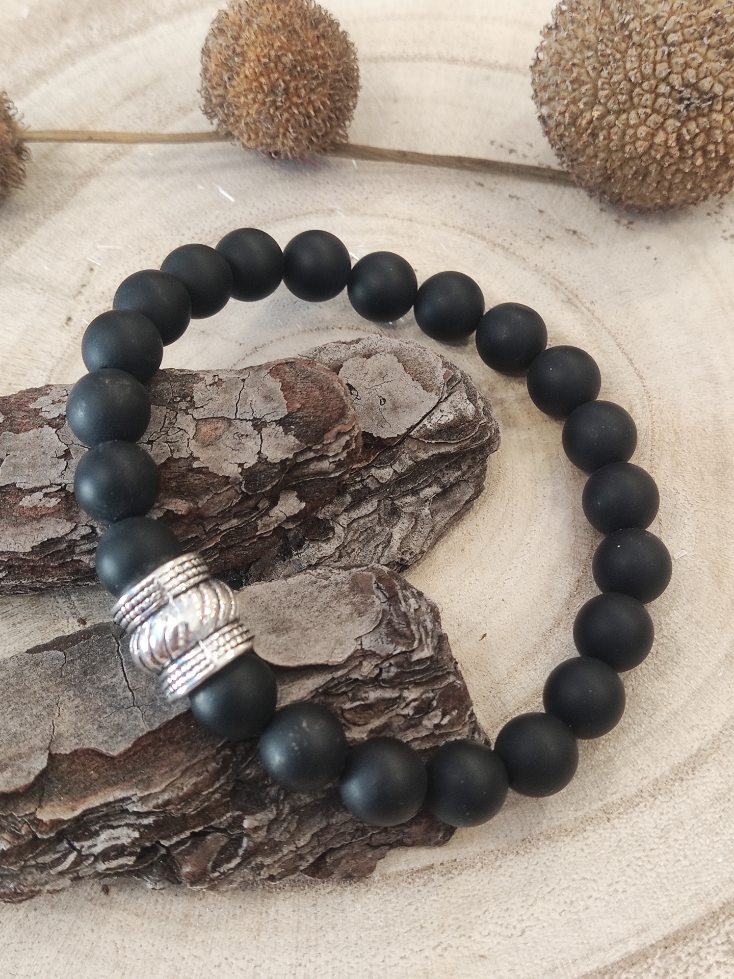 Pulseira preta fosca com detalhe metálico prateado sobre madeira clara e elementos decorativos naturais